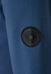 Blauwe gestructureerde stof met een cirkelvormige zwarte logopatch. De textuur heeft een ribbelpatroon, waardoor de visuele diepte en interesse wordt versterkt.
