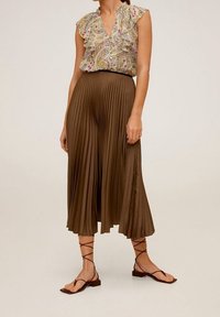 Blumige Bluse mit Rüschensäumen, kombiniert mit einem langen, gefalteten braunen Rock. Das Outfit wird durch flache Sandalen mit Riemen ergänzt.