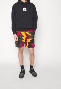 Uomo con felpa nera con cappuccio, pantaloncini con motivo zigzag vivaci di colore giallo, rosso e nero, calzini neri e scarpe da ginnastica nere, in piedi su sfondo bianco.