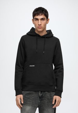 Młody mężczyzna ubrany w czarną bluzę z kapturem z logo "Jack & Jones" i wyblakłe czarne dżinsy, stojący na jednolitym białym tle.