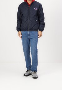 Mann in einer marineblauen Steppjacke mit Reißverschluss und blauen Jeans, der vor einem einfarbigen weißen Hintergrund steht. Graue Sneaker sind sichtbar.