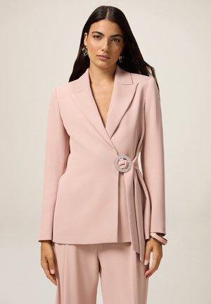 Blazer ajusté rose pâle avec revers cranté, cintré à la taille avec un broche en strass rond et des manches longues.