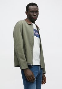 Κυπαρισσί πράσινο μπουφάν με καφέ γιακά από κορντουρέ, ελαφρύ ύφασμα, ίσια γραμμή. Φορεμένο πάνω από λευκό μπλουζάκι με στάμπα και μπλε τζιν.