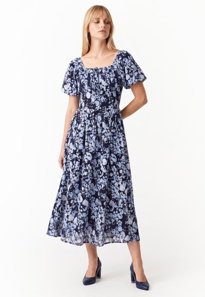 Donna che indossa un vestito midi blu navy con stampa floreale azzurra e maniche a sbuffo, abbinato a scarpe con tacco alto blu navy.