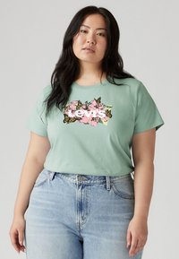 Mint green t-shirt with a floral and butterfly graphic in pink and yellow. Short sleeves and relaxed fit, paired with light blue denim jeans.