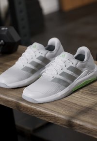 Paire de chaussures de course Adidas blanches et grises avec semelles vertes posées sur une surface en bois, haltère en arrière-plan flou.