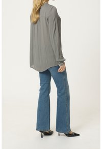 Blouse grise légère à manches longues avec texture plissée ; assortie à un jean bleu évasé et des escarpins noirs à bout pointu.