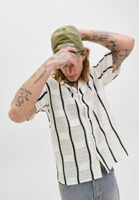 Chemise tricotée blanche à manches courtes avec des rayures noires et des motifs texturés. Casquette vert olive avec texte blanc. Tatouages visibles sur les bras.