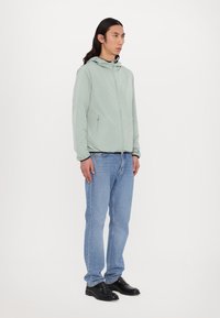 Lys grønt, hættejakke med lynlås foran og sidelommer, parret med blå denimjeans og sorte sko. Minimalistisk design.