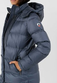 Giubbotto imbottito blu navy con cappuccio, dotato di chiusura con zip, tasche laterali e logo sulla manica sinistra. Texture liscia e lucida.