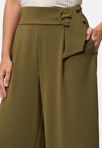 Olive green wide-leg trousers with a textured finish, featuring a front tie belt with a circular accent and side pockets.