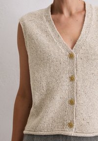 Gestrickte ärmellose Weste in Beige mit V-Ausschnitt und fünf braunen Knöpfen vorne. Verfügt über einen strukturierten Stoff und eine leicht lockere Passform.
