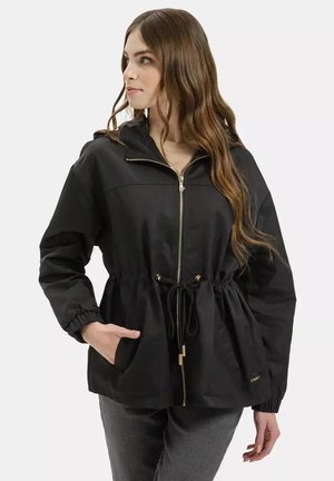 Młoda kobieta z długimi włosami w czarnej kurtce na zamek z regulowanym paskiem w talii i elastycznymi mankietami, stojąca z jedną ręką w kieszeni.