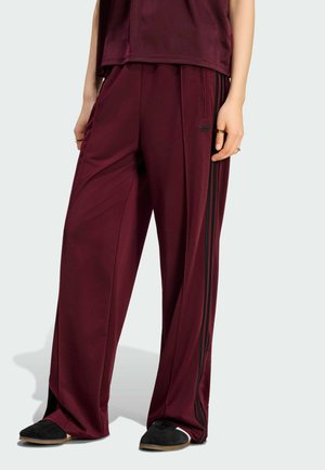 Persona che indossa pantaloni della tuta bordeaux a gamba larga con strisce laterali nere e scarpe slip-on nere, in piedi davanti a uno sfondo semplice.