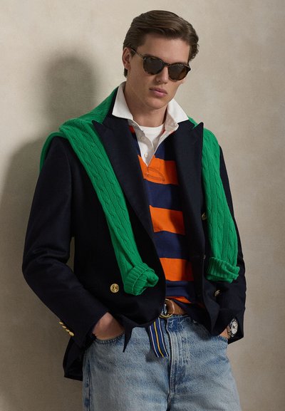 Marineblazer mit goldenen Knöpfen, grüner Kabelstrickpullover lässig über die Schultern gelegt, orange- und marineblau gestreiftes Poloshirt, helle Jeans aus Denim, Sonnenbrille.