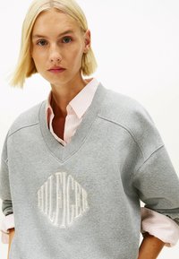 Felpa grigia in cotone con scollo a V, con "HILFIGER" ricamato in bianco. Indossata sopra una camicia colletto a righe rosa con maniche arrotolate.