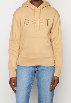 Person wearing beige GANT hoodie with front pocket and drawstrings paired with light blue denim jeans, standing against a plain background.