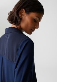 Marineblaue Bluse mit einem glatten, satinähnlichen oberen Abschnitt und einem strukturierten unteren Abschnitt, ausgestattet mit einem klassischen Kragen und langen Ärmeln.