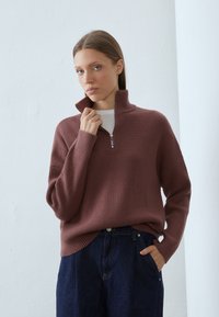Žebrovaný svetr tmavě mauve s vysokým límcem, polo-zipovým zapínáním a spadlými rameny, v kombinaci s tmavě modrými džíny.