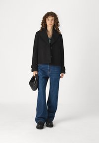 Black cropped jacket with a wide collar, two buttons, and rolled sleeves, paired with flared blue jeans and a black handbag.