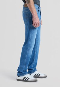 Mann trägt blaue Lee-Jeans und weiße Adidas-Sneakers und steht seitlich vor einem einfarbigen Hintergrund.