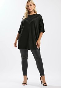 Kvinna står klädd i en lös svart topp med spetsdetaljer i chevronmönster, svarta läderleggings och svarta högklackade sandaler mot en vit bakgrund.