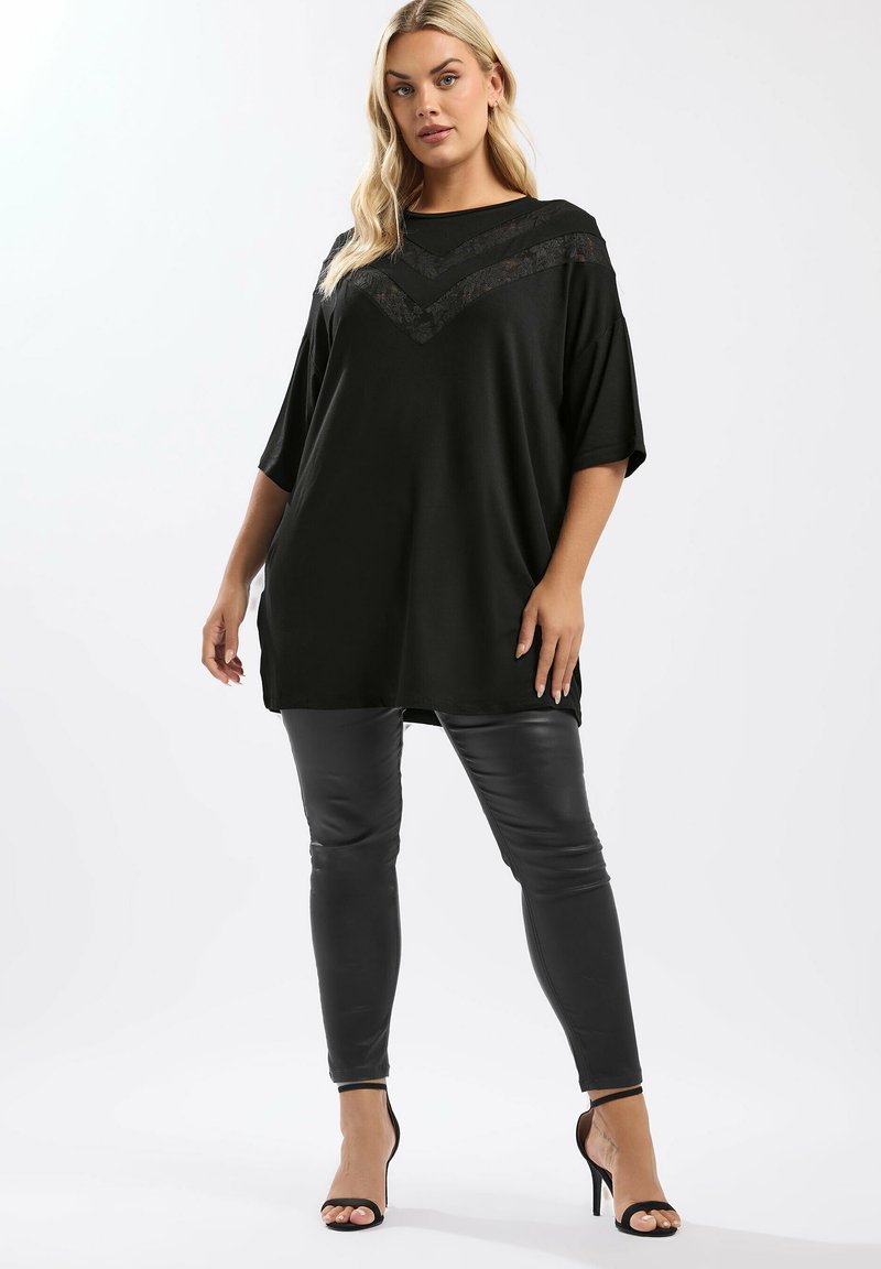 Kvinna står klädd i en lös svart topp med spetsdetaljer i chevronmönster, svarta läderleggings och svarta högklackade sandaler mot en vit bakgrund.