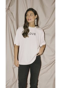 Volná růžová tričko s nápisem "LOVE" černým písmem, v kombinaci s tmavými džíny. Modelka má dlouhé vlnité vlasy a visící náušnice.