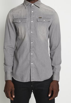 Man draagt een vervaagde grijze blouse met knopen en twee borstzakken, en zwarte broek, handen ontspannen langs het lichaam tegen een effen achtergrond.