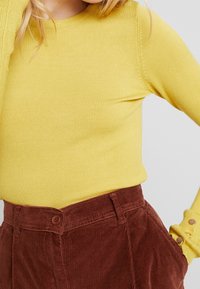 Žlutý priléhavý pletený svetr s žebrovanou texturou, doplněný hnědými manšestrovými kalhotami s vysokým pasem, které mají knoflíkový detail a boční kapsy.