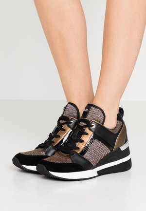 Czarne i brązowe metaliczne sneakersy z białymi podeszwami noszone na bosych stopach, z sznurowadłami i sportowym klinowym obcasem.