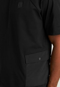 Maglietta nera a maniche corte in cotone con taschino a patta dotato di bottone a pressione, design minimale e un branding discreto sul petto.