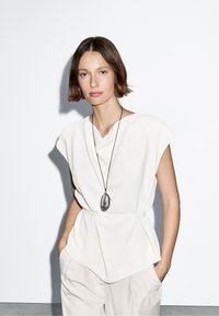 Hvid, kortærmet bluse med draperet halsudskæring og peplum-kant, båret over lys beige, løs pasform bukser; har en lang, sort pendul halskæde.