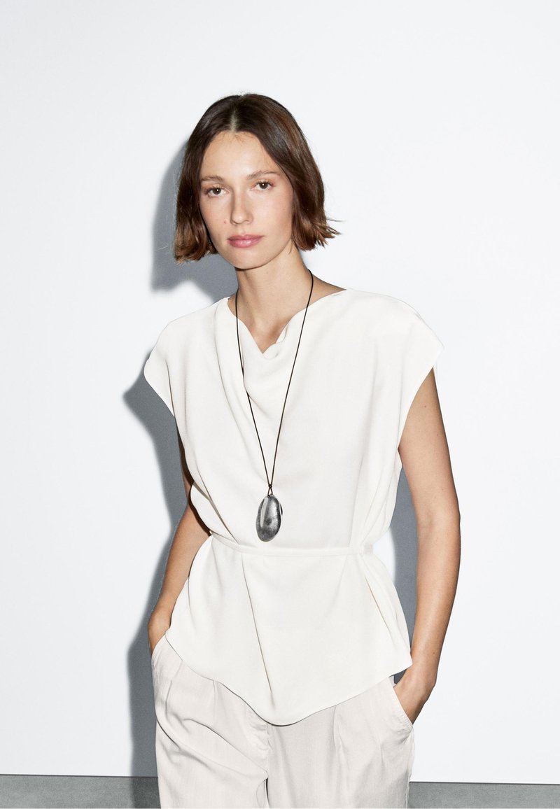 Hvid, kortærmet bluse med draperet halsudskæring og peplum-kant, båret over lys beige, løs pasform bukser; har en lang, sort pendul halskæde.