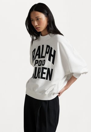 Biała bluza z dużym czarnym napisem "RALPH LAUREN" z przodu, okrągły dekolt i luźne rękawy. Czarne spodnie noszone na dole.