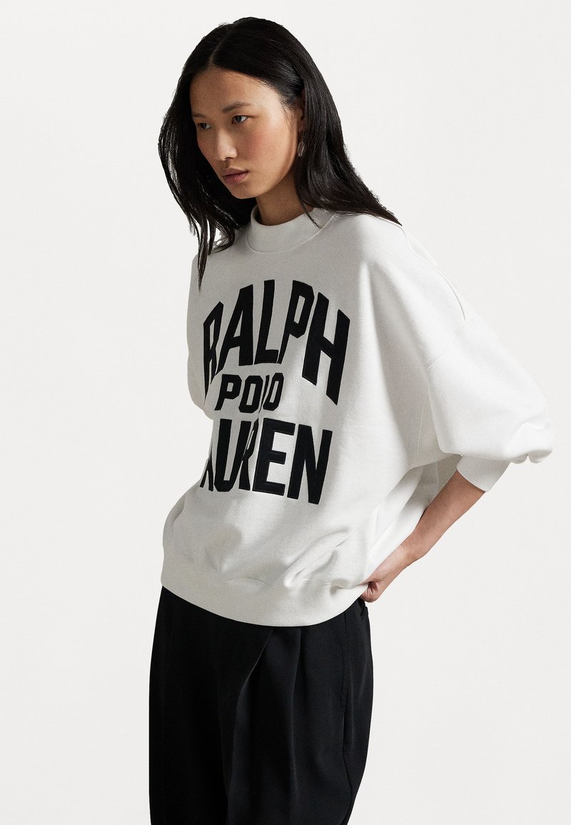 Biela mikina s výrazným čiernym nápisom "RALPH LAUREN" na prednej strane, okrúhly výstrih a voľné rukávy. Pod ňou sú oblečené čierne nohavice.