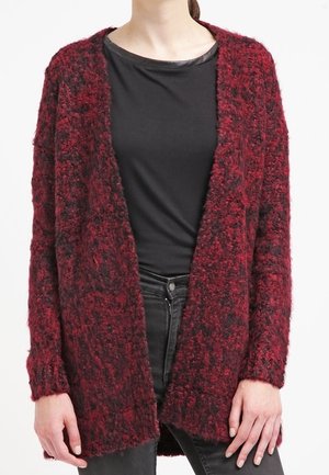 Kobieta ubrana w teksturowany, otwarty sweter z długim rękawem w kolorze ciemnoczerwonym i czarnym, nałożony na czarną koszulę i czarne spodnie.