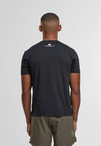 Muž s krátkými vlasy, oblečený v černém tričku s malým bílým textem na horní části zad a olivově zelených šortkách, stojící na jednolitém pozadí.