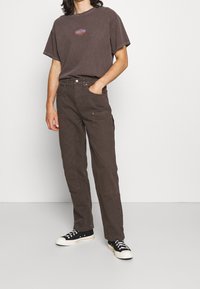 Hnědé oversized tričko s barevným potiskem, doplněné hnědými taktovými kalhotami s několika kapsami a černými teniskami s bílými podrážkami.