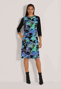 Vrouw in een knielang zwart jurkje met blauw en groen bloemenpatroon, driekwart mouwen, zwarte enkelboots, staande met één hand in de zak.