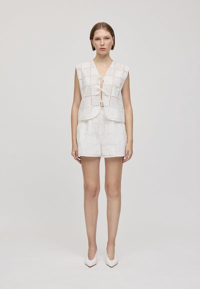 Haut sans manches blanc avec design découpé et détail à nouer, associé à un short assorti ; texture lisse, style minimaliste. Chaussures : talons blancs.