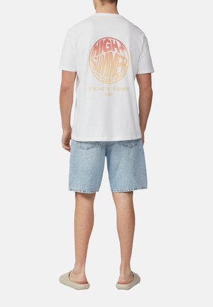Weißes Baumwoll-T-Shirt mit einem runden, pinken und orangen Grafikdesign auf der Rückseite, das Text und Koordinaten zeigt. Getragen mit hellblauen Denim-Shorts.