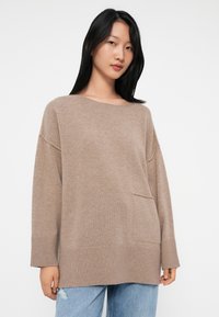 Beżowy sweter z dzianiny o szerokim dekolcie, opadających ramionach, przedniej kieszeni oraz ściągaczach na rękawach i dolnym brzegu, w połączeniu z niebieskimi dżinsami.