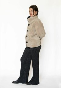 Beige pelsjakke i imiteret pels med sorte knapper i togglestil, høj krave og løs pasform, kombineret med sorte glitrende vidde bukser og sorte sko.