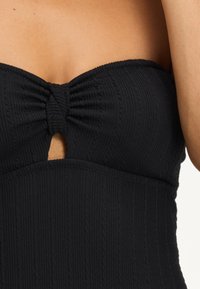 Schwarzer Bandeau-Badeanzug mit strukturiertem Stoff, ausgestattet mit einer gedrehten Mitte und einem kleinen Ausschnitt. Das Design weist gerippte Details über das Material auf.