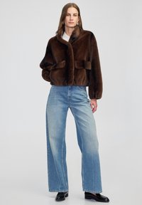 Braune Kunstfelljacke mit Schalkragen, zwei Fronttaschen und einer cropped Passform, kombiniert mit hellblauen, weit geschnittenen Jeans und schwarzen Schuhen.