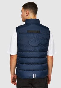 Marine puffer gilet met hoge kraag, zichtbare stiksels en een reliëf ontwerp van een esdoornblad op de achterkant. Heeft een zwart label en twee witte strepen aan de onderkant.