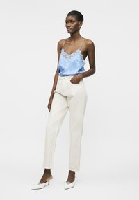 Femme portant un caraco en satin bleu clair bordé de dentelle, un pantalon taille haute écru et des talons pointus blancs, debout avec une main dans la poche.