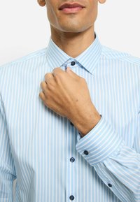 Hellblau gestreiftes Hemd mit Knopfkragen. Es verfügt über schmale weiße Streifen, blaue Knöpfe und eine strukturierte Stoffoberfläche für einen gepflegten Look.