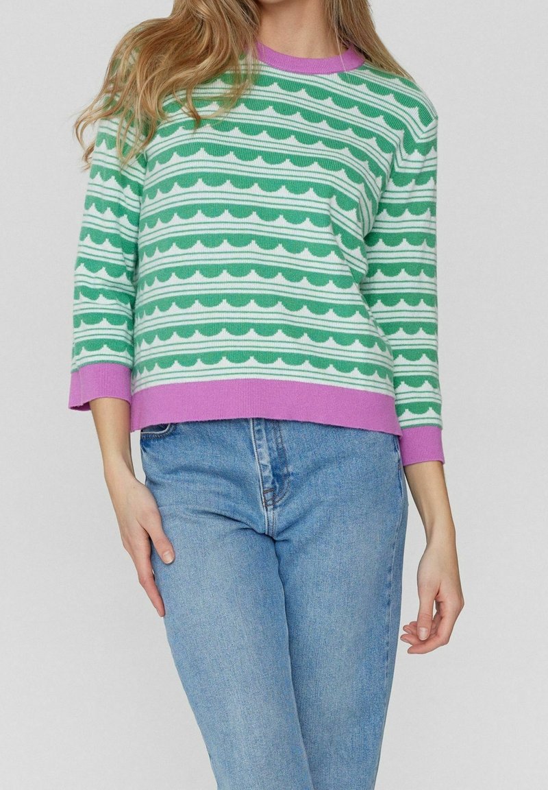 Pull-over rayé vert et blanc avec motifs ondulés, texture côtelée, col rond violet et manches trois-quarts. Porté avec un jean bleu.